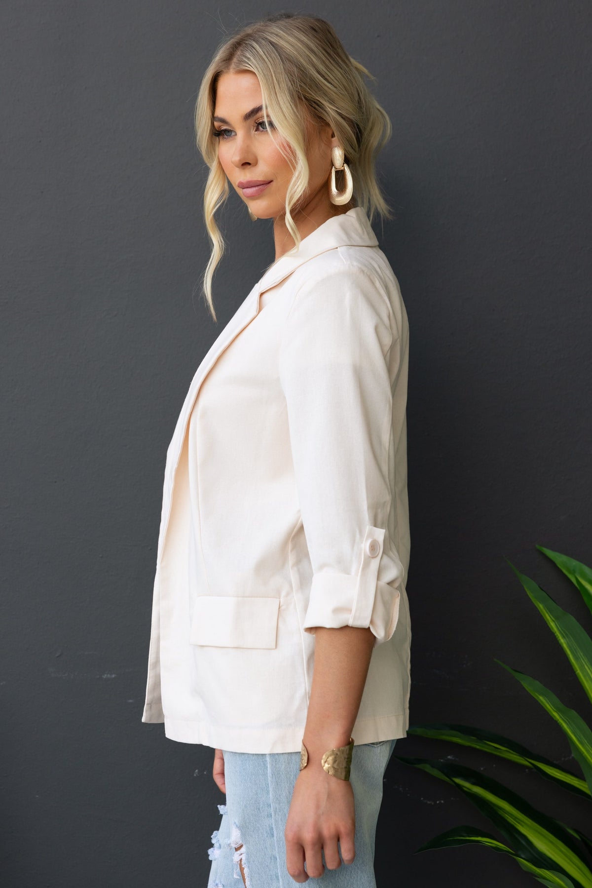 Model wearing the Merci Blazer in beige, featuring a collar, lapels, and faux pockets, set against a dark background.