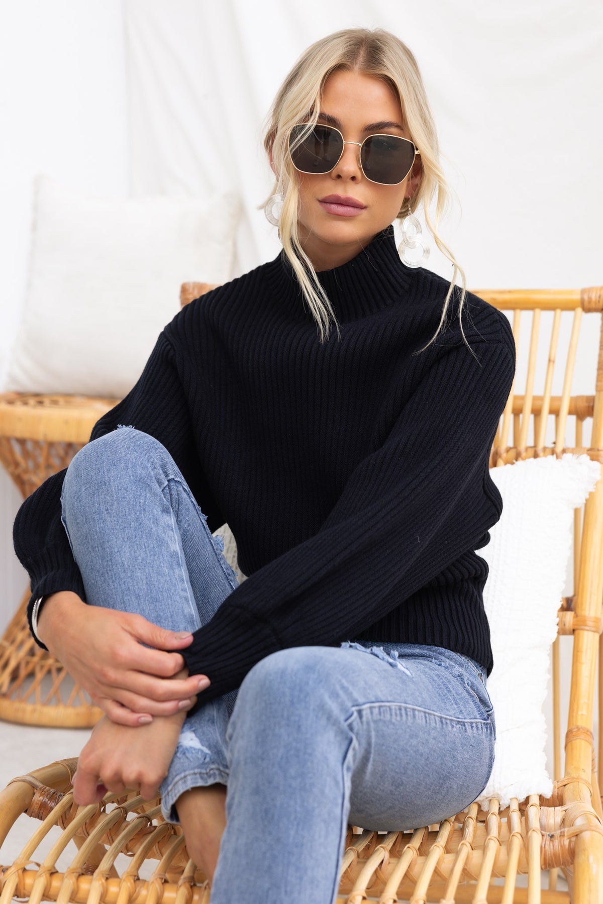 Model wearing a black ribbed Sola Jumper with mock neckline and wide sleeves, paired with jeans and sunglasses.