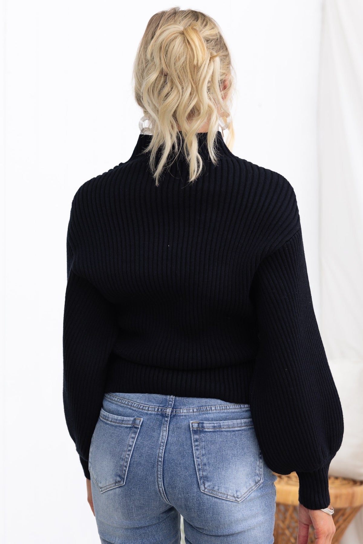 Back view of a stylish black ribbed Sola jumper with mock neckline and wide sleeves, paired with blue jeans.