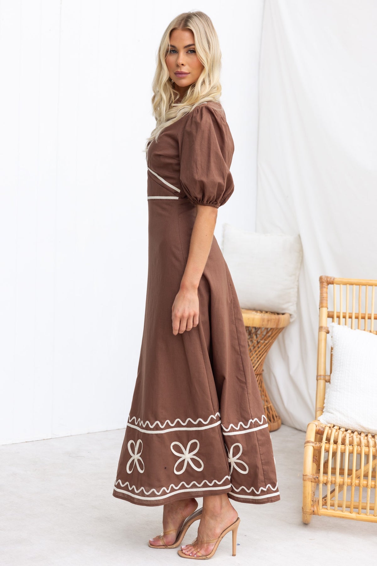 Model wearing a brown Azurra maxi dress with puff sleeves and floral embroidery, styled in a minimalist setting.