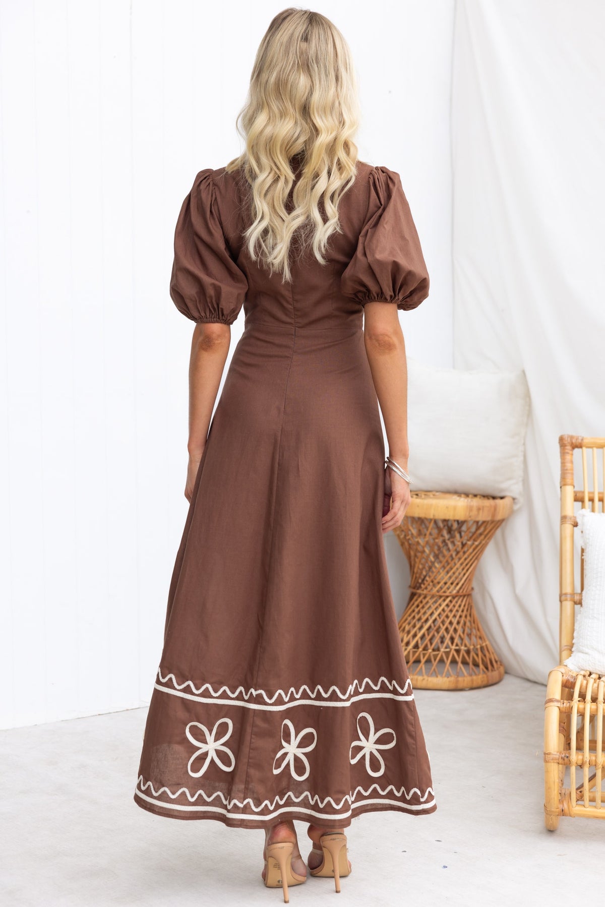 Model wearing the Azurra Maxi Dress in brown, showcasing a back view with puff sleeves and floral embroidery at the hem.