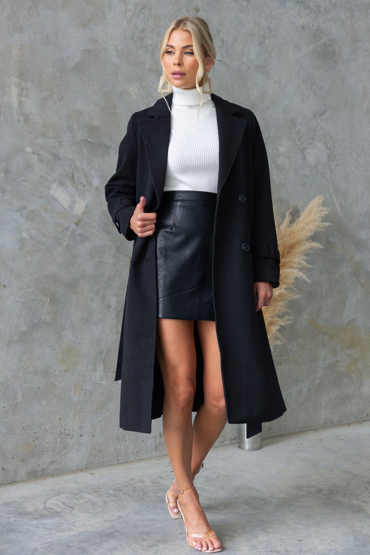 Model wearing Indah Skirt, a black faux leather mini skirt, paired with a stylish long coat and white turtleneck.