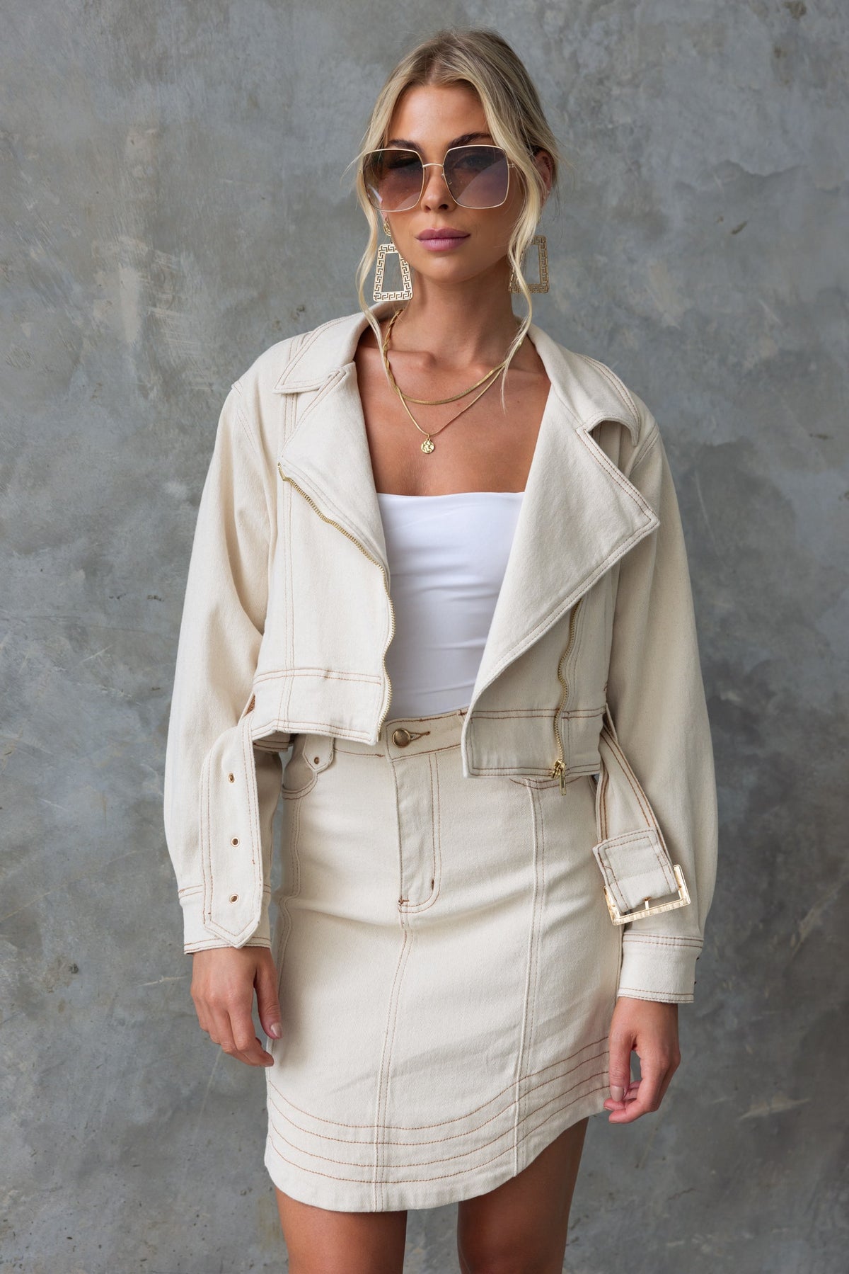 Model wearing a cream colored denim jacket with a zip-up front, paired with a matching skirt and sunglasses.