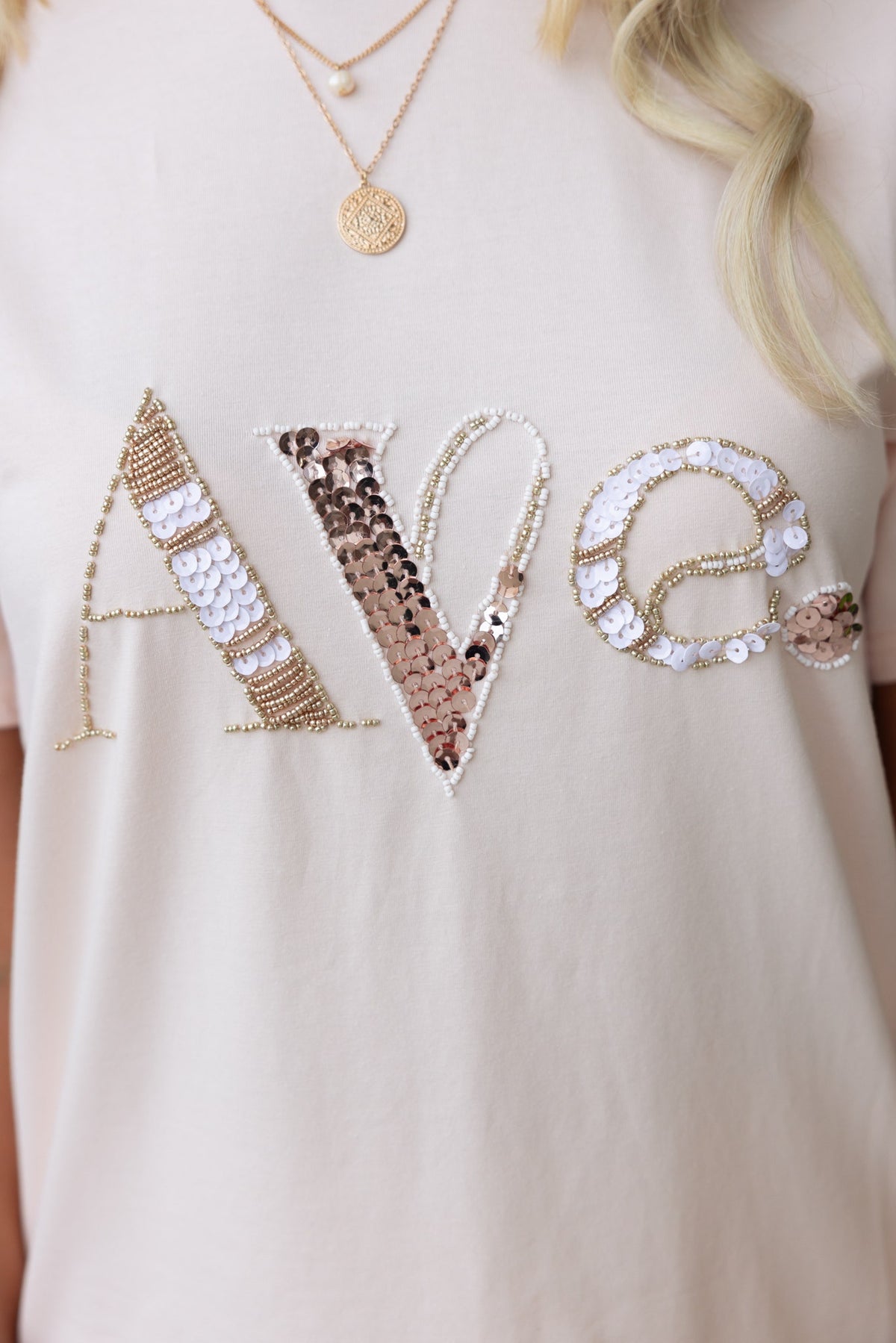 Close-up of the Cesca Tee with embellished 'Ave' lettering in sequins and beads, showcasing a round neckline and soft cotton texture.
