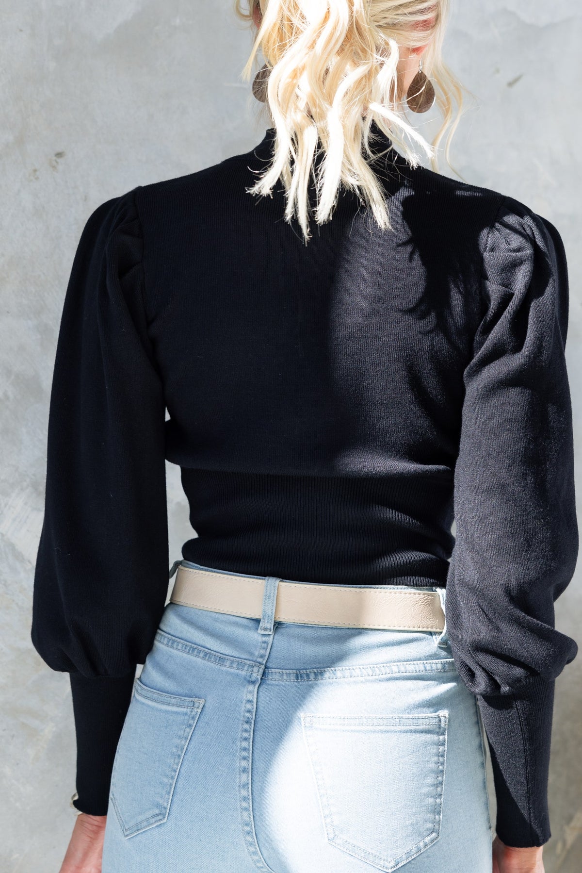 Back view of Sania Top, a black knit top featuring puffed sleeves and a high neckline, styled with light blue jeans.