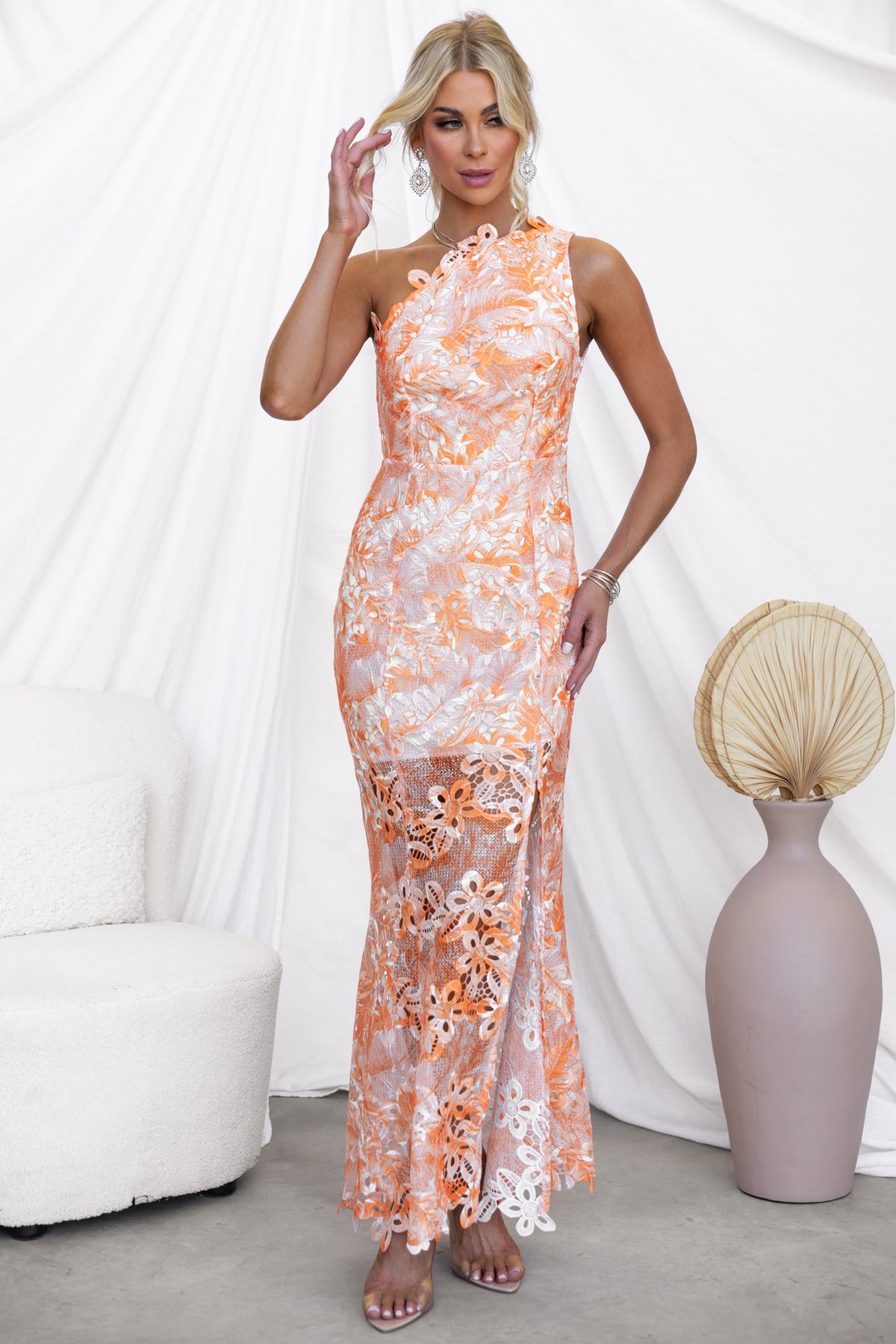Model wearing a vibrant orange and white one-shoulder Racha Maxi Dress with floral design and a front split.