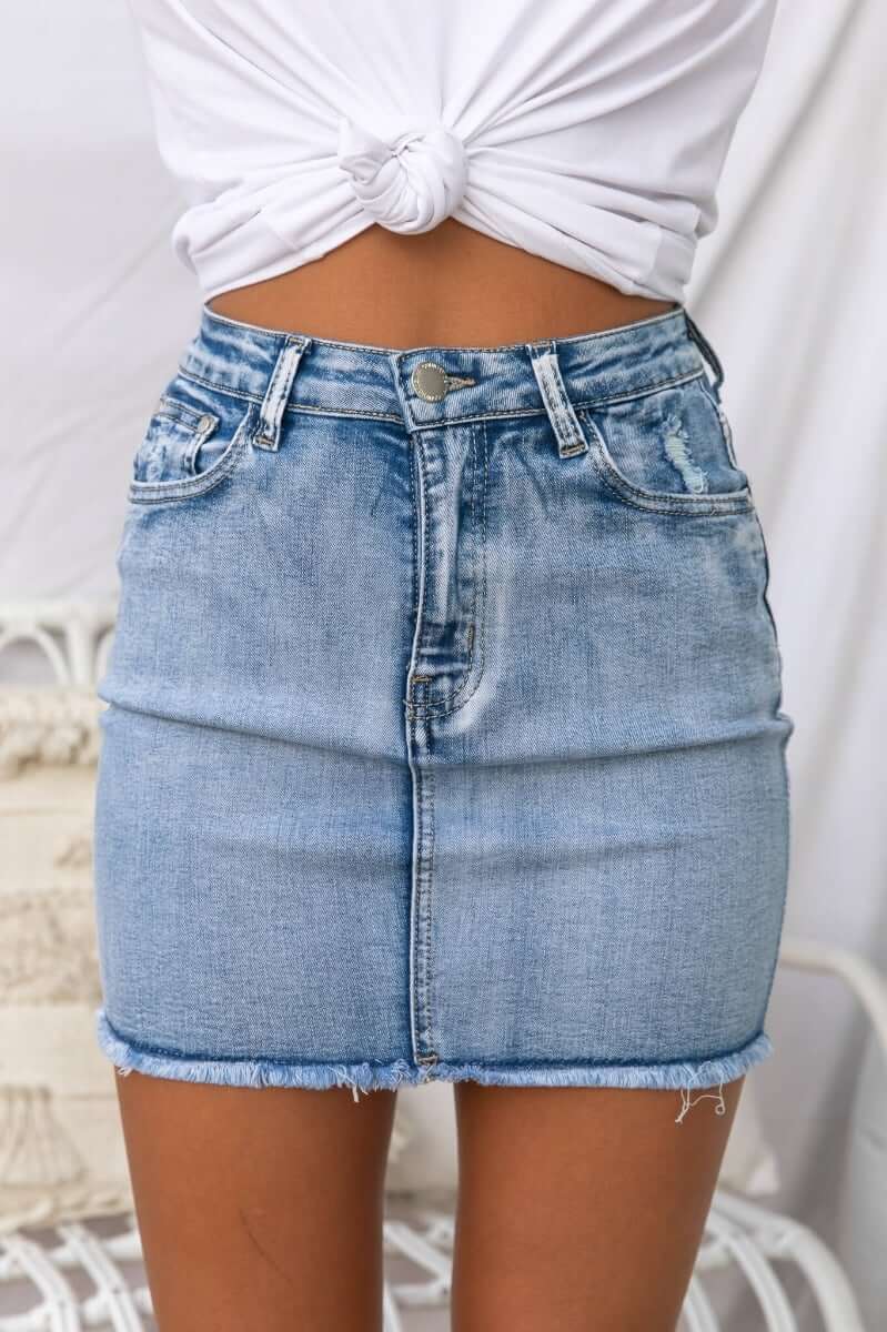 Light blue denim skirt with distressed details and raw hemline, styled with a knotted white top.