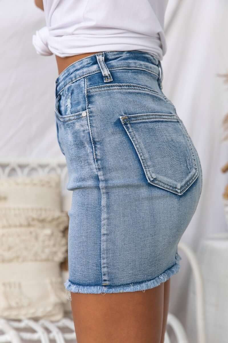Side view of Nice For What Skirt featuring distressed pockets, raw hemline, and a standard fit in light blue denim.