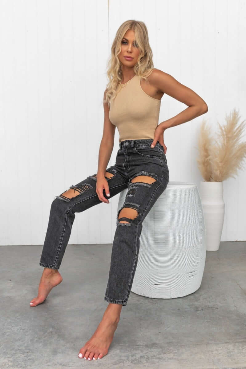 Model wearing black wash Pasha Mom Jeans with distressed rips, paired with a beige top and styled in a modern setting.