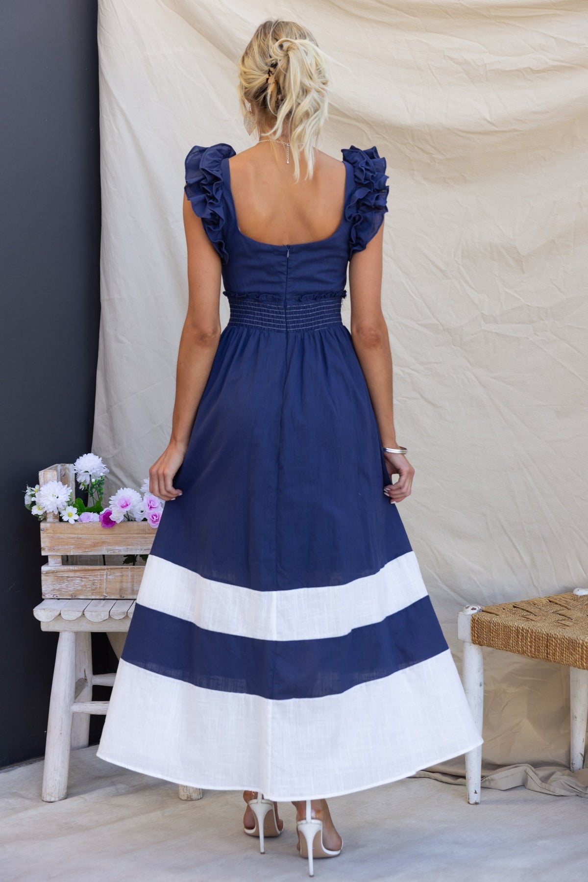 Back view of Kape Maxi Dress with navy fabric, ruffled sleeves, and white stripes at the hem, styled on a model.