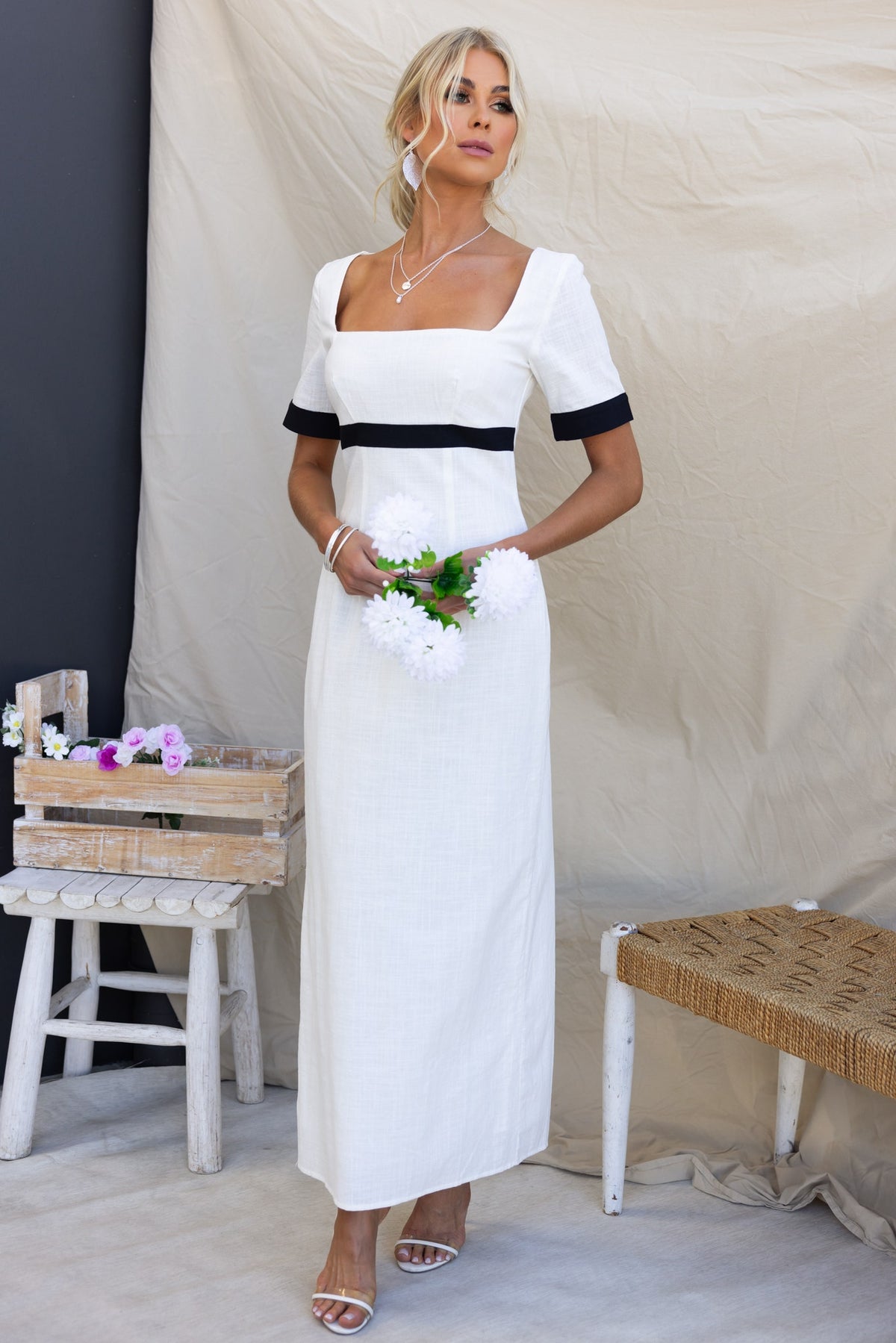 Soria Maxi Dress featuring a square neckline, back split, and linen-like texture, styled with a bouquet of flowers.