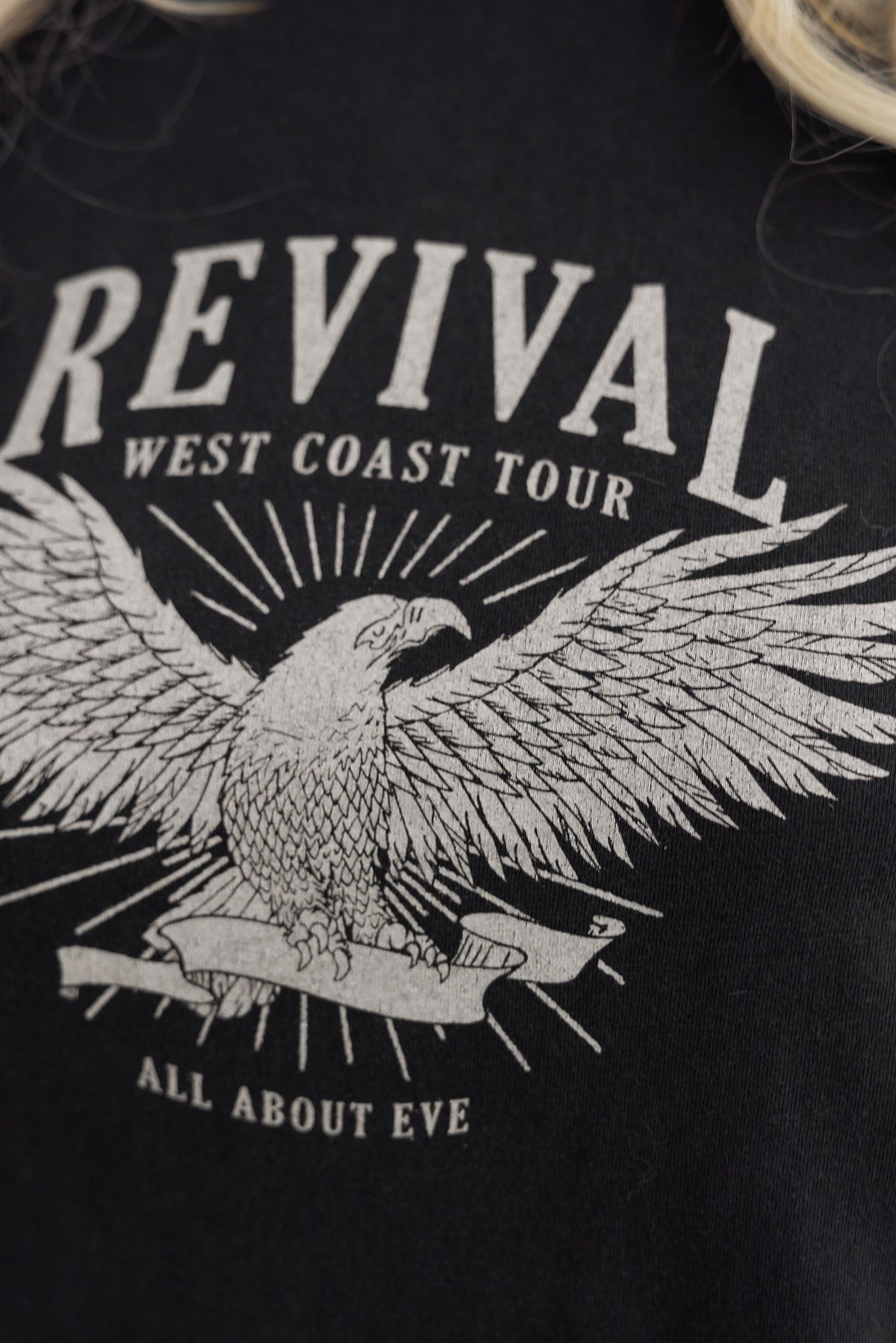 Close-up of black t-shirt featuring "Revival West Coast Tour" graphic with eagle, part of All About Eve collection.