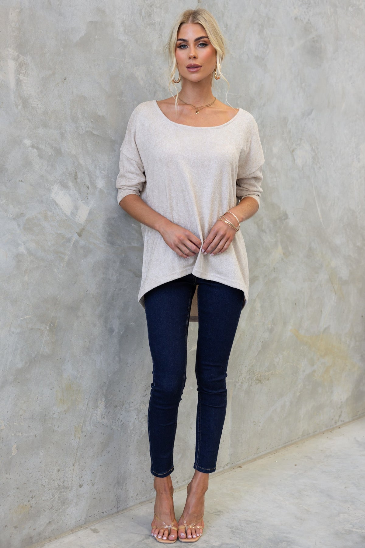 Model wearing Vargo Top with Boat neckline and high-low hem, styled with dark jeans and sandals against textured backdrop.