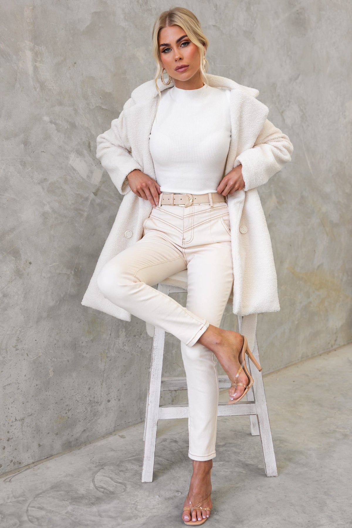 Woman in a stylish neutral outfit featuring a tawny coat, fitted white top, and light jeans. Elegant footwear completes the look.