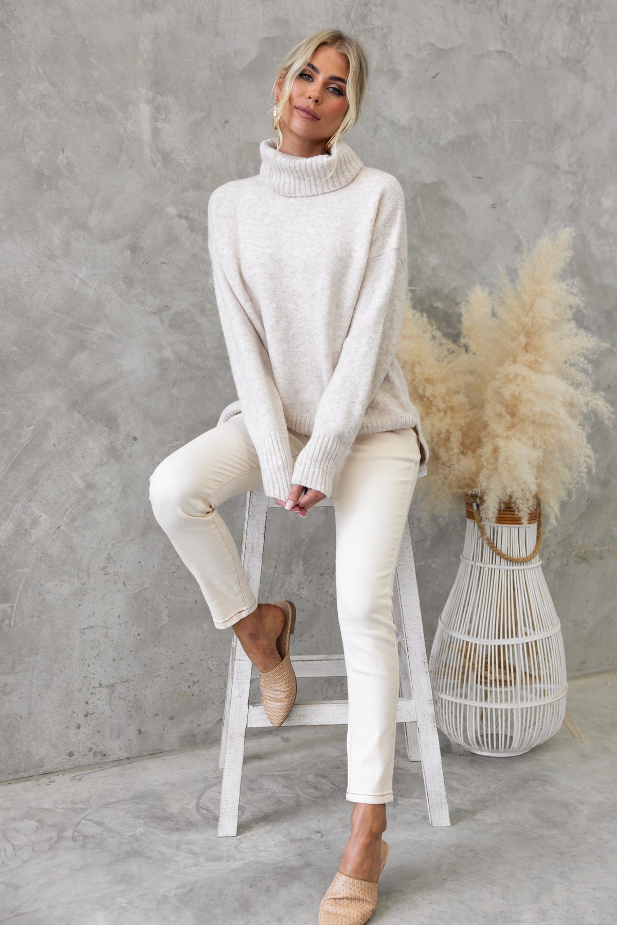 Woman wearing a cozy cream Orion Jumper with a turtle neckline, styled with white pants and sitting on a stool.