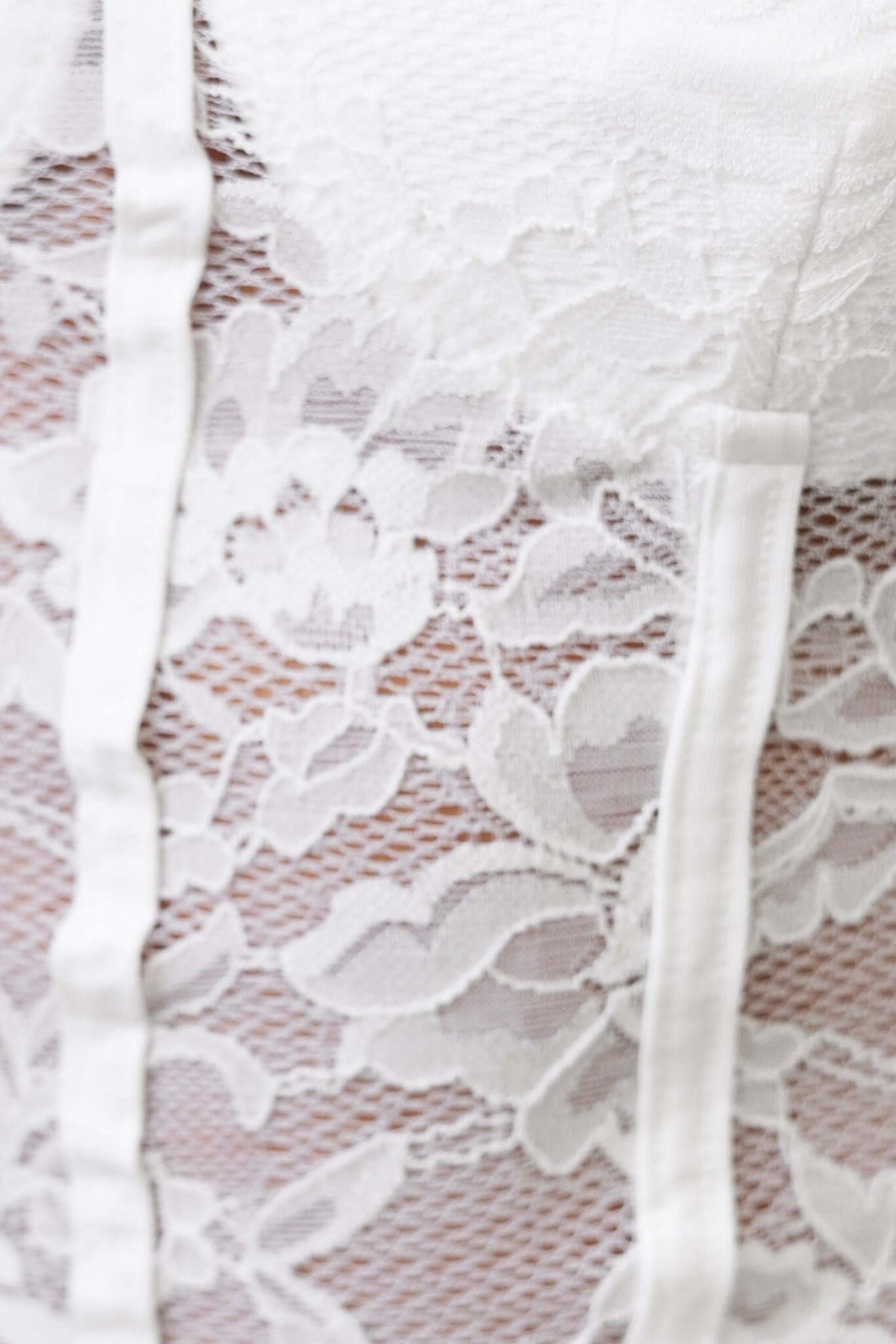 Close-up of white lace bodysuit with adjustable straps and faux boning, showcasing intricate floral design.