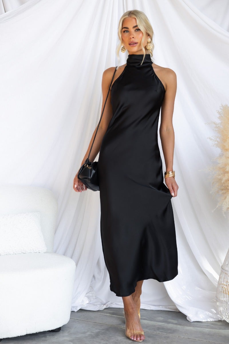 Model wearing a black satin Humble Maxi Dress with a high tie neckline, posing in a stylish setting.