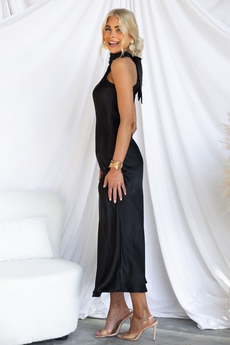 Model wearing a black satin maxi dress with a high tie neckline and side zip, featured in a stylish indoor setting.