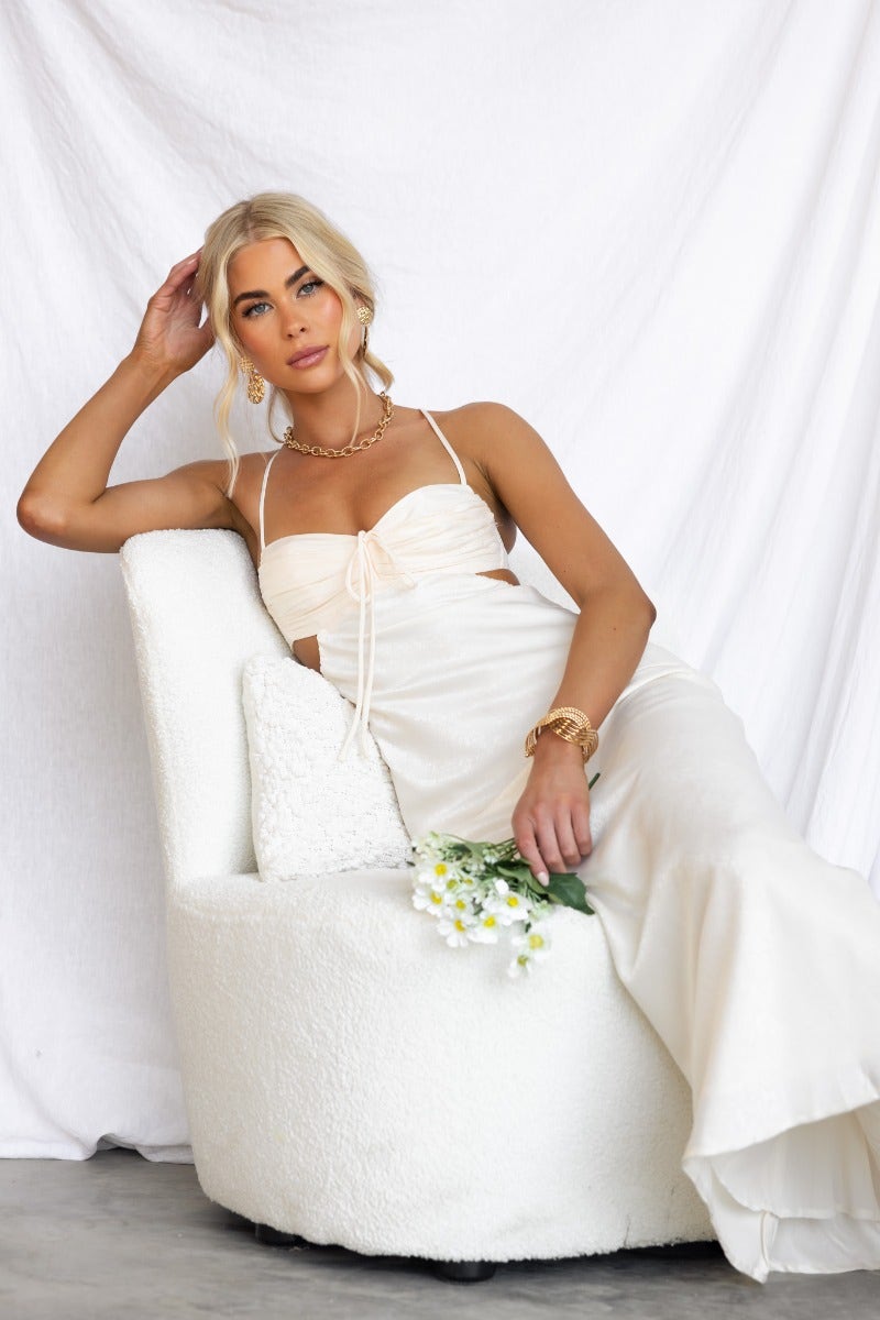 Model wearing Bayside Maxi Dress with adjustable straps and tie detail, sitting on a white chair, holding flowers.
