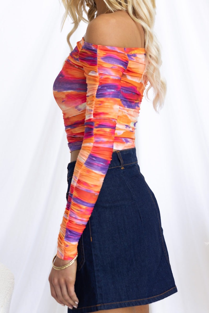 Off-shoulder mesh overlay Elsie Top in vibrant colors, paired with a denim skirt, showcasing ruching and a semi-sheer look.