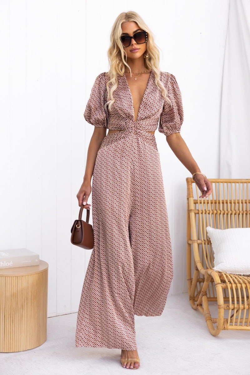 Model wearing the Soho Jumpsuit in satin with V-neckline, puff sleeves, and cut-out waist, styled with sunglasses.