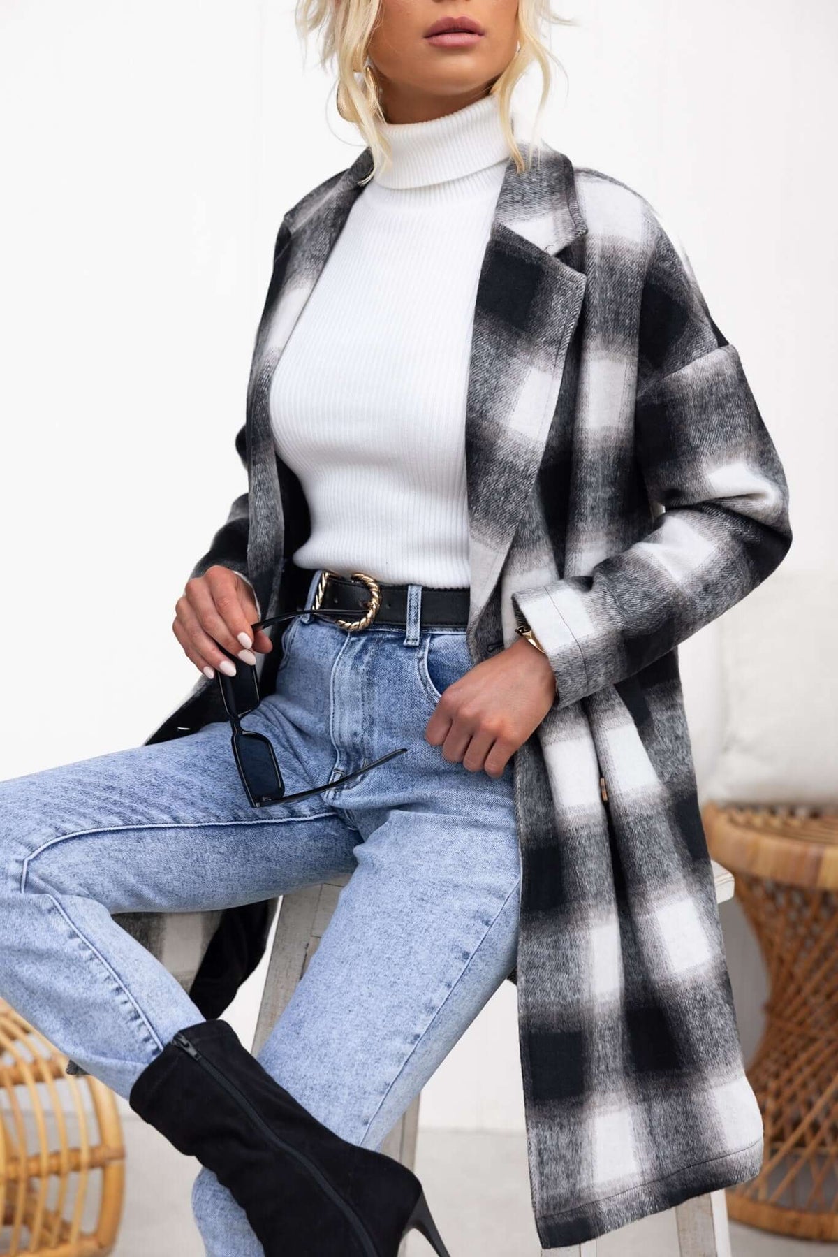 A woman wearing a black and white plaid Tiffin coat over a white turtleneck and blue jeans, styled with a belt.