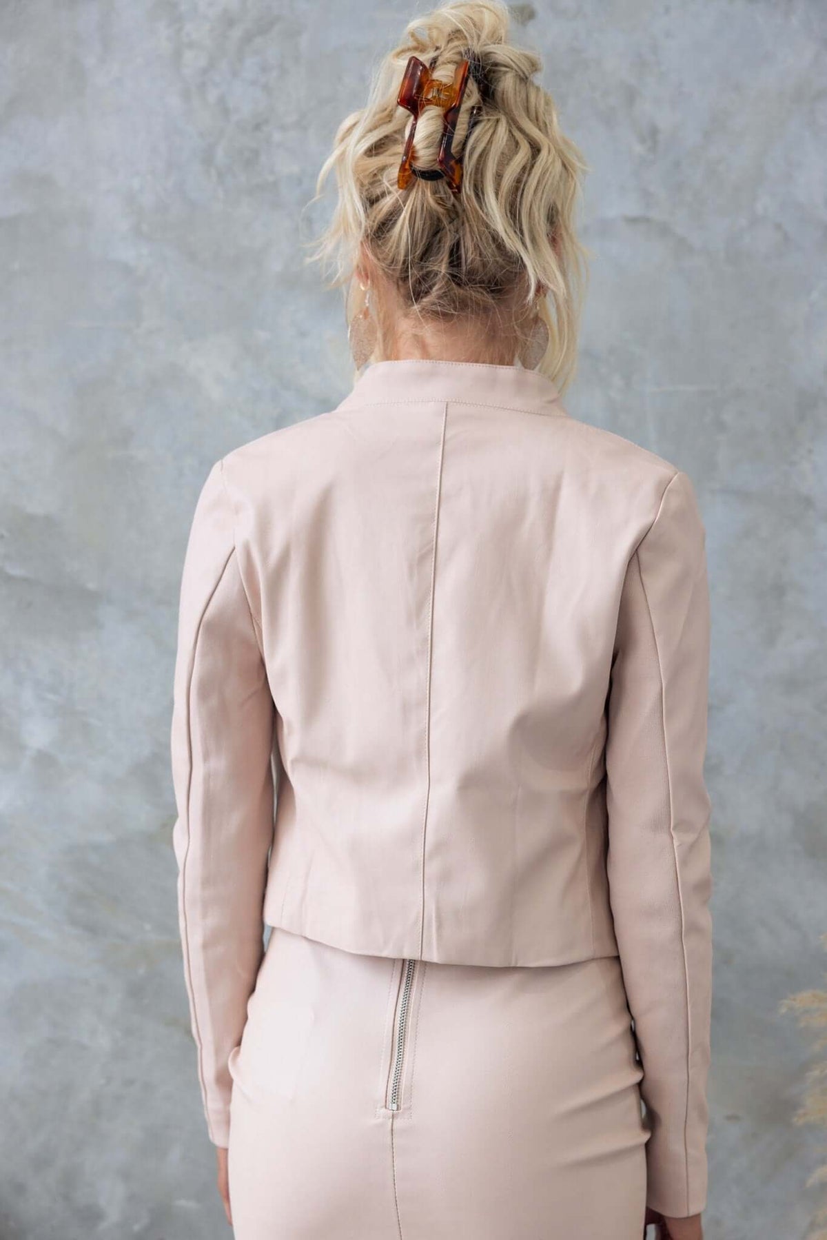 Back view of a woman wearing the Udine Blazer, a fitted faux leather jacket in soft pink, styled with hair bows.