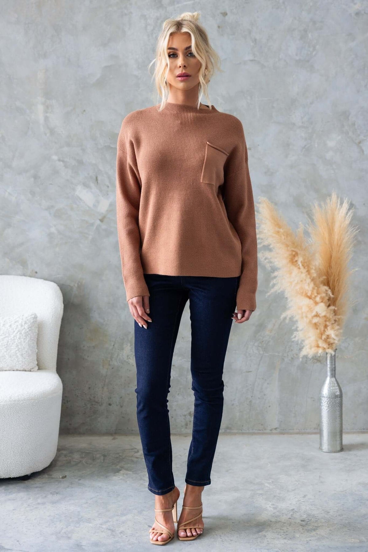 Model wearing a brown Palladia Jumper with a round neckline and pocket, paired with dark jeans against a neutral backdrop.