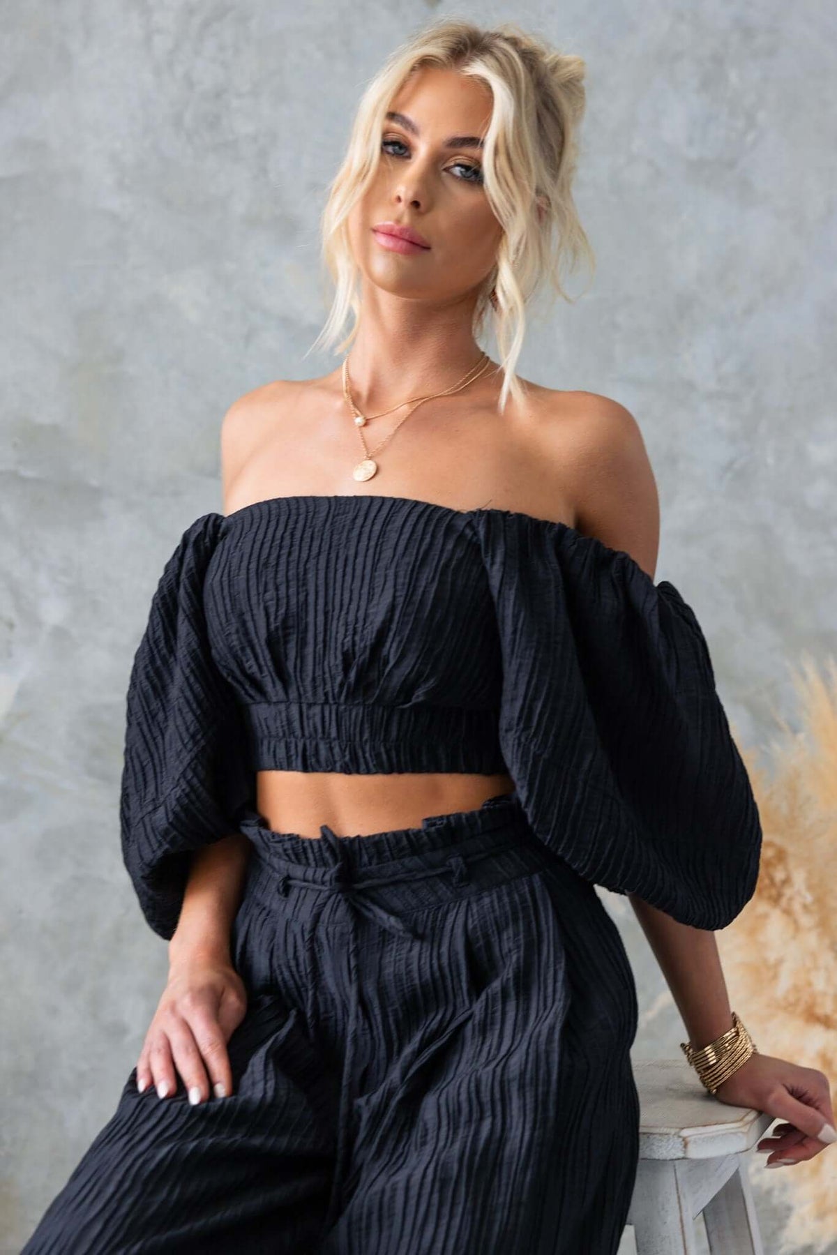 Model wearing a stylish black off-shoulder top with textured material and wide sleeves, paired with matching pants.
