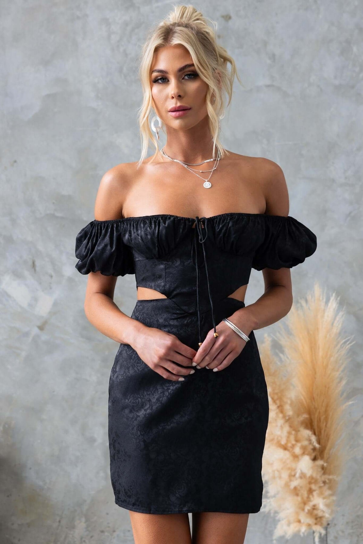 Stylish Dalma Dress in black with off-shoulder sleeves, cut-out waist, and satin finish for a chic look.