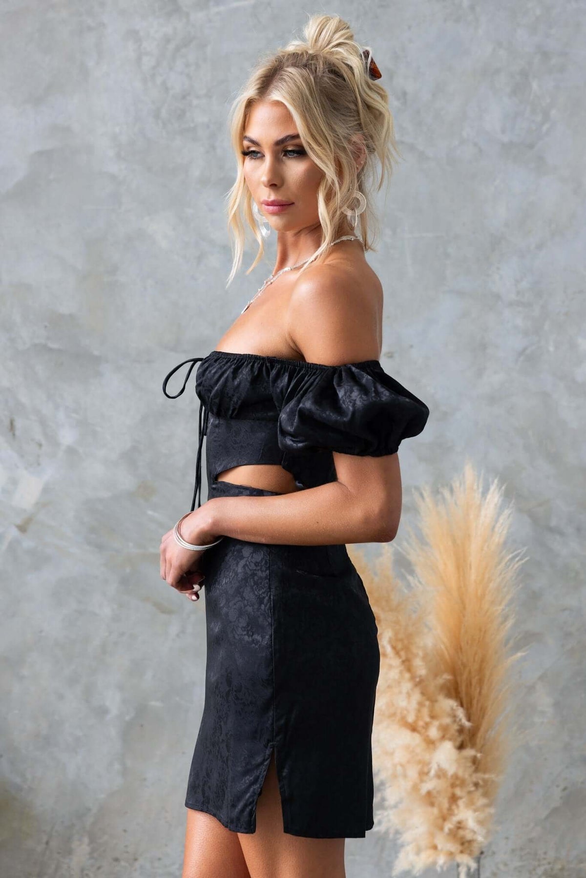 Model wearing a black Dalma Dress with puff sleeves, cut-out waist, and front split against a textured backdrop.