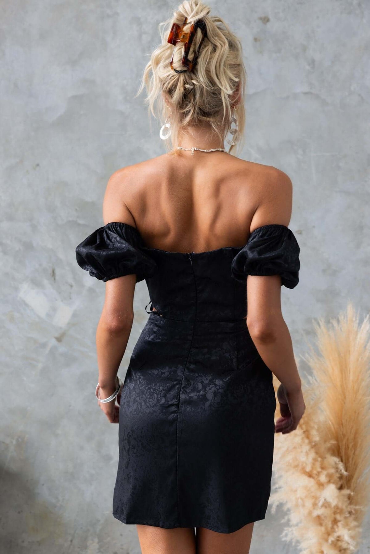 Back view of Dalma Dress, a black satin look dress with puff sleeves and a cut-out waist.
