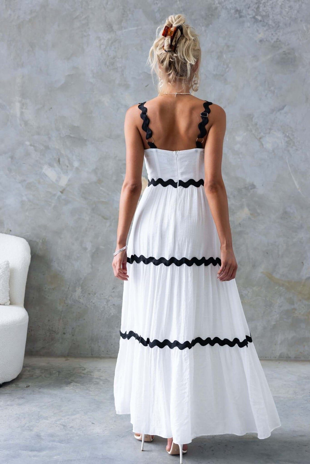 Back view of the Qaia Maxi Dress in white with adjustable straps and ruffled black accents against a textured background.