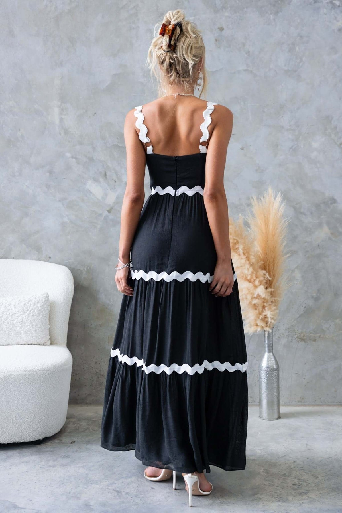 Back view of a woman wearing a black Qaia maxi dress with adjustable straps and white scalloped trim.