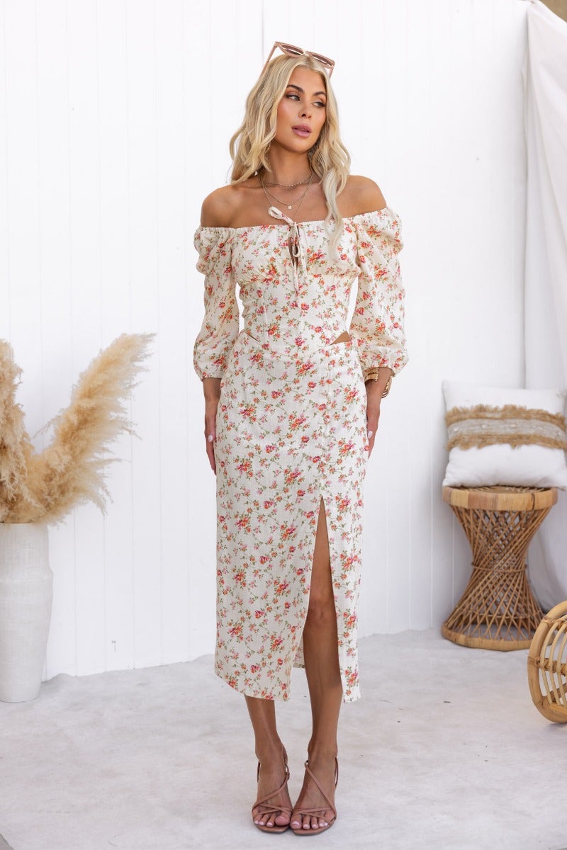 Model wearing a floral off-the-shoulder dress with puff sleeves, featuring a tie detail and side slit, styled with sandals.