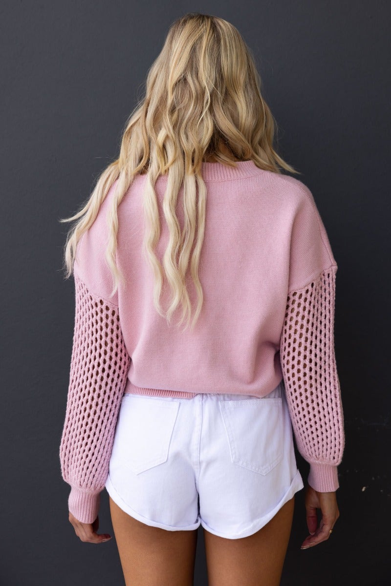Back view of a model wearing a pink Verny jumper with open weave sleeves, paired with white shorts.