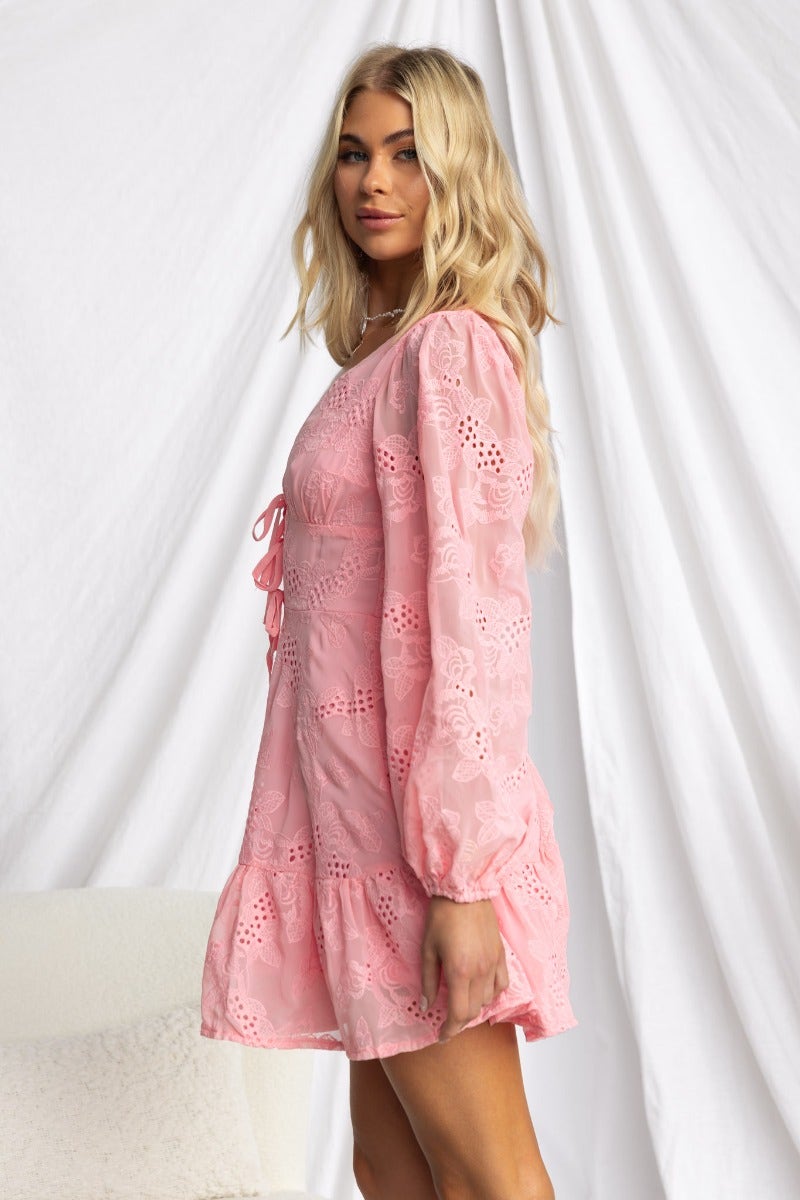 Model wearing a pink Anglaise dress with V-neckline and tie detail, featuring elastic cuffs and a flared skirt.
