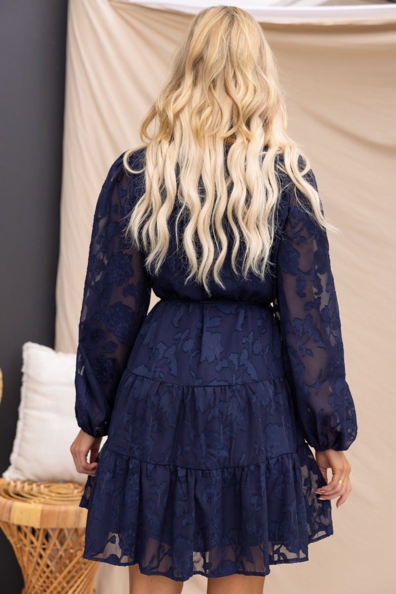 Back view of a woman wearing a navy embossed print Halston dress with elastic sleeves and a tiered skirt.