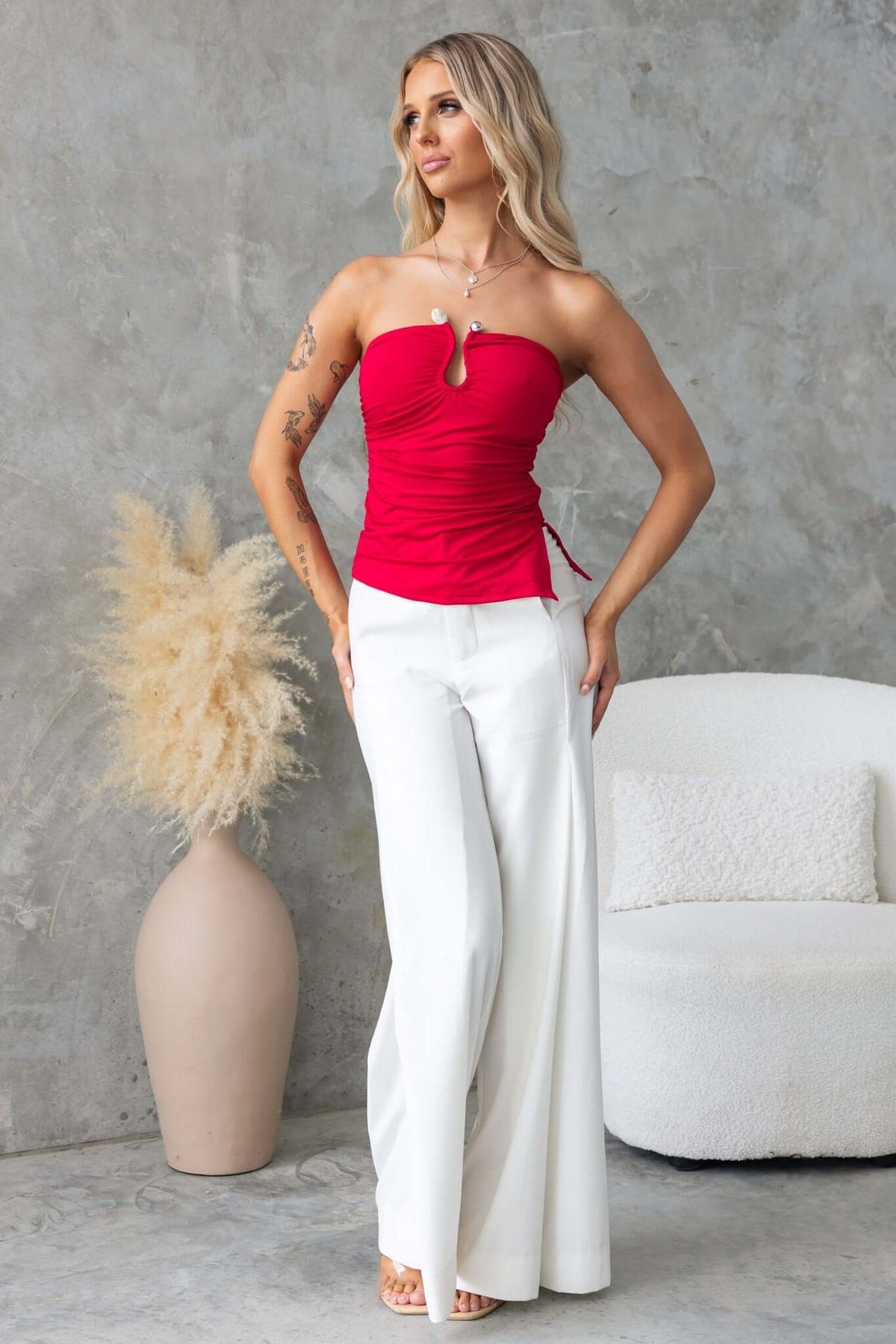 Model wearing a red strapless Kylie Top with silver hardware detailing, paired with white wide-leg pants.