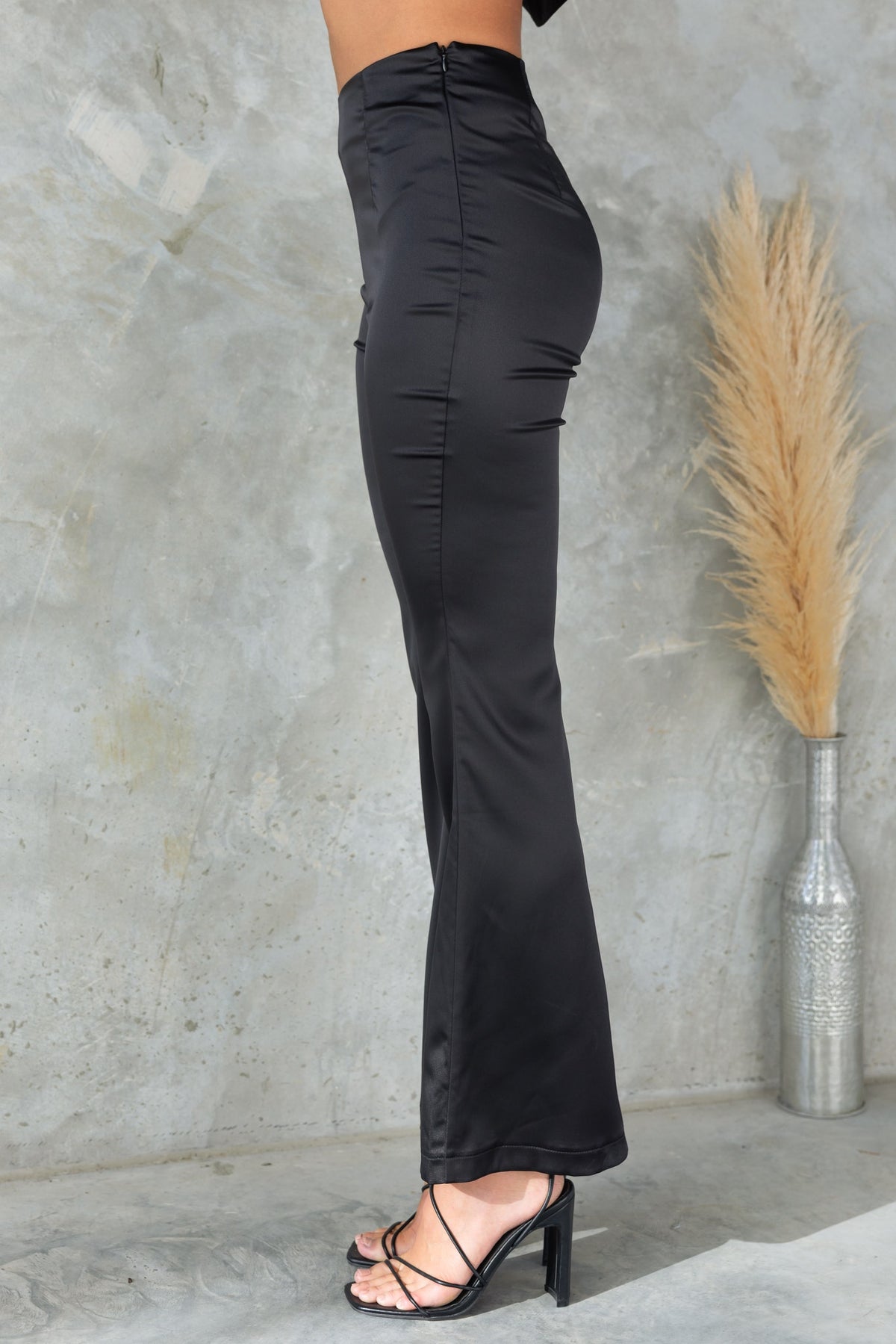 Amore Pants in black, featuring mid-high waist, flare leg, and satin finish, styled with heels against a modern background.