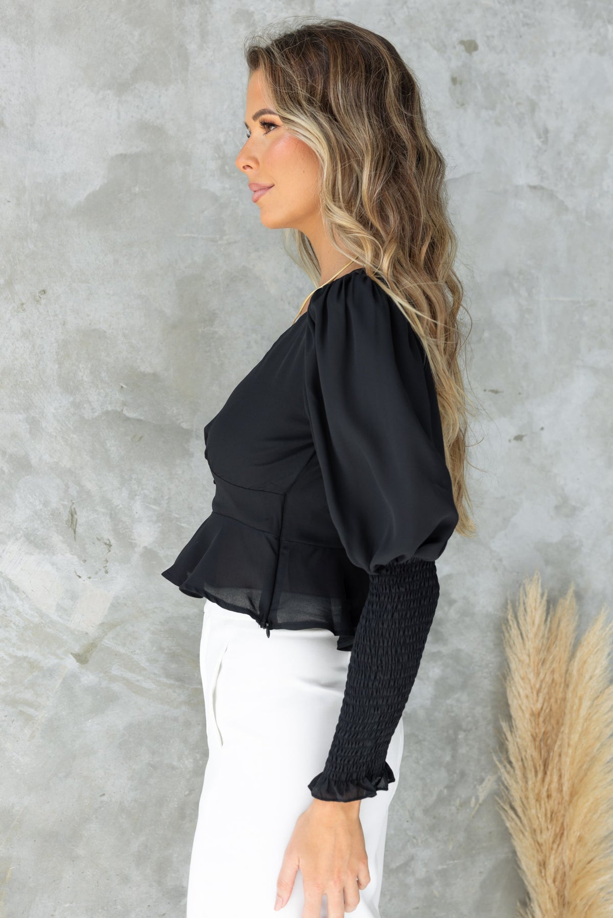 Side view of a model wearing a black Fleur Top with puff sleeves and a peplum hem, styled with white pants.