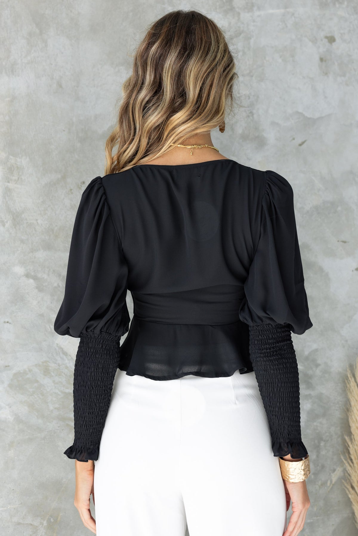 Back view of the Fleur Top featuring puff sleeves and smocked detailing in black, styled with white pants.