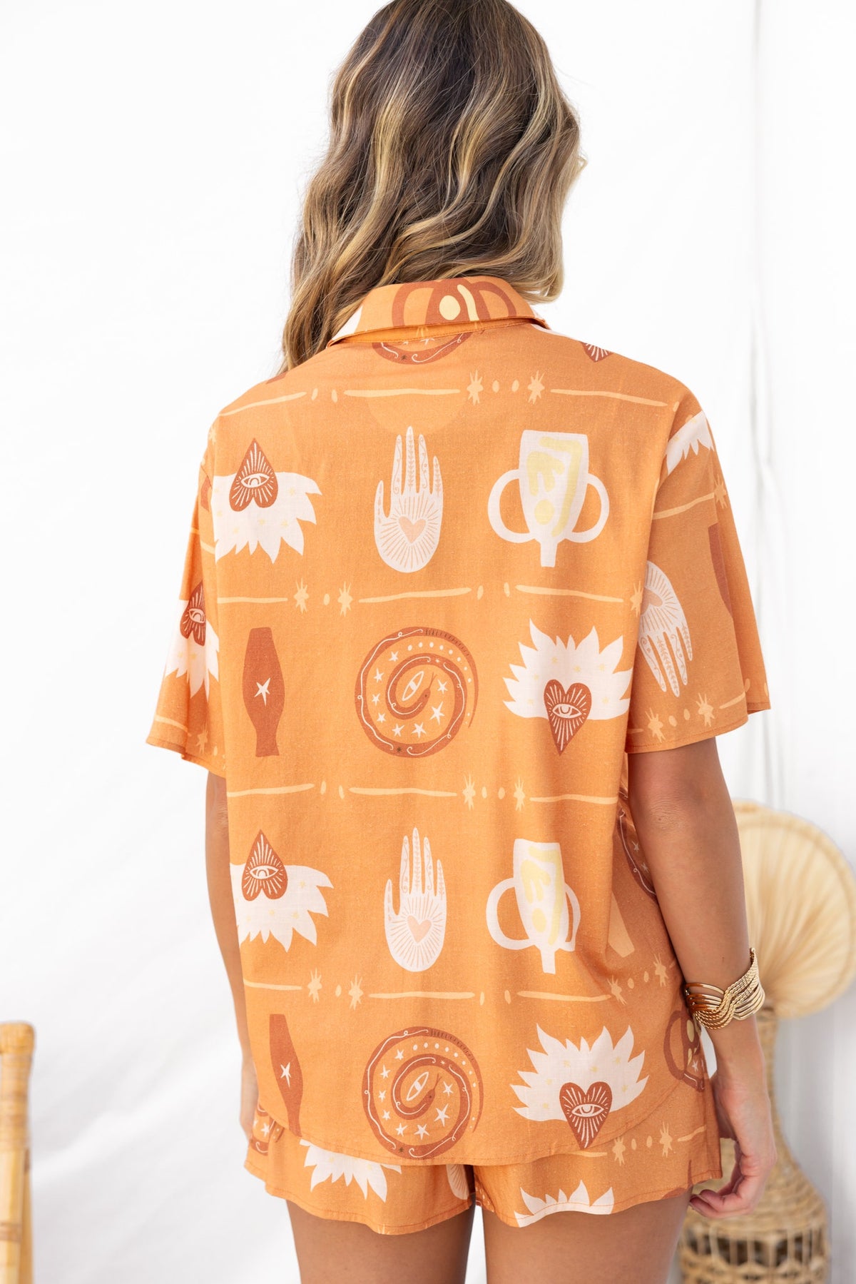 Back view of a model wearing an orange patterned Koffie set with shorts and a button-up shirt.