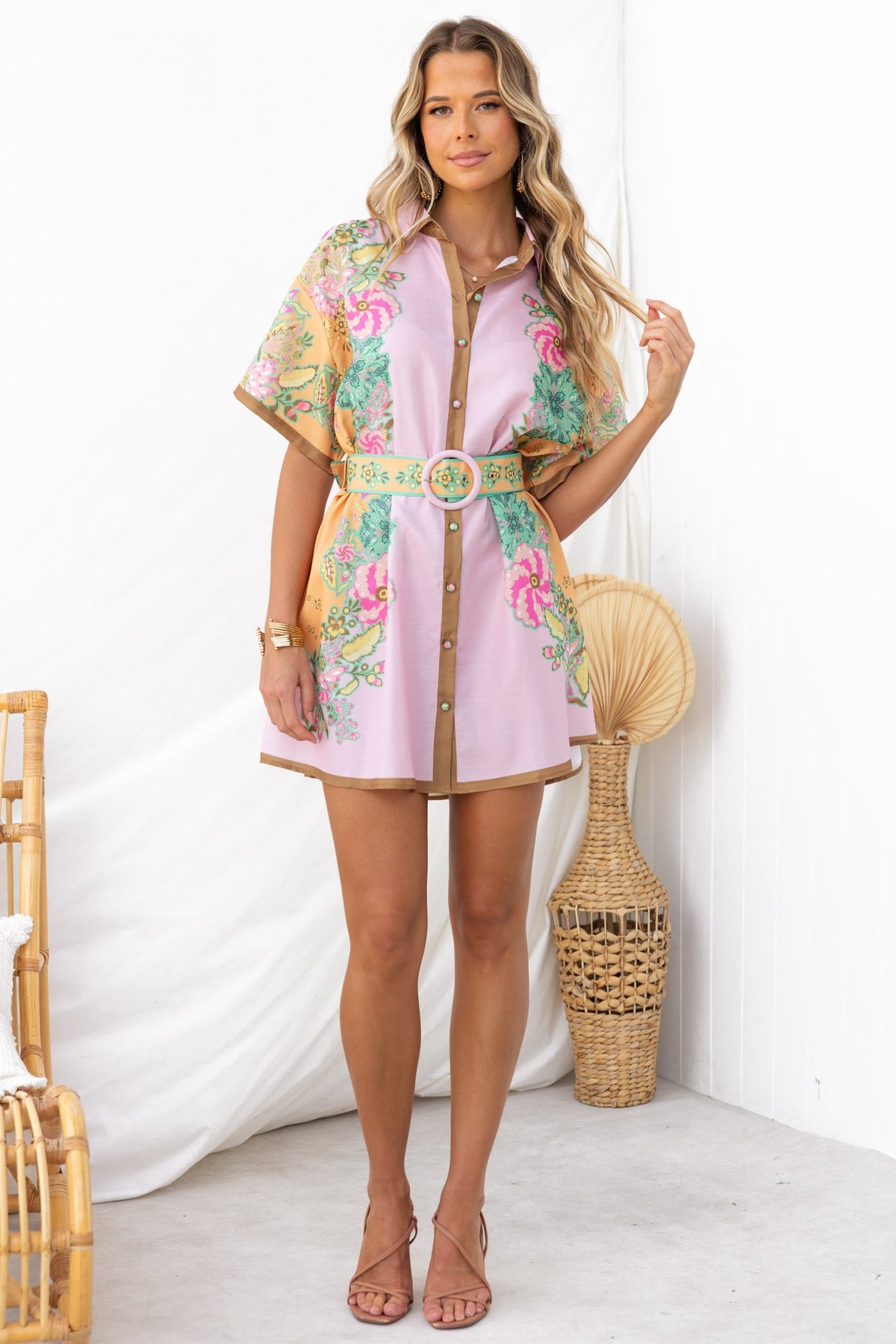 Model wearing Jaziel Dress, a pink button-up dress with floral pattern and removable belt, in a stylish indoor setting.