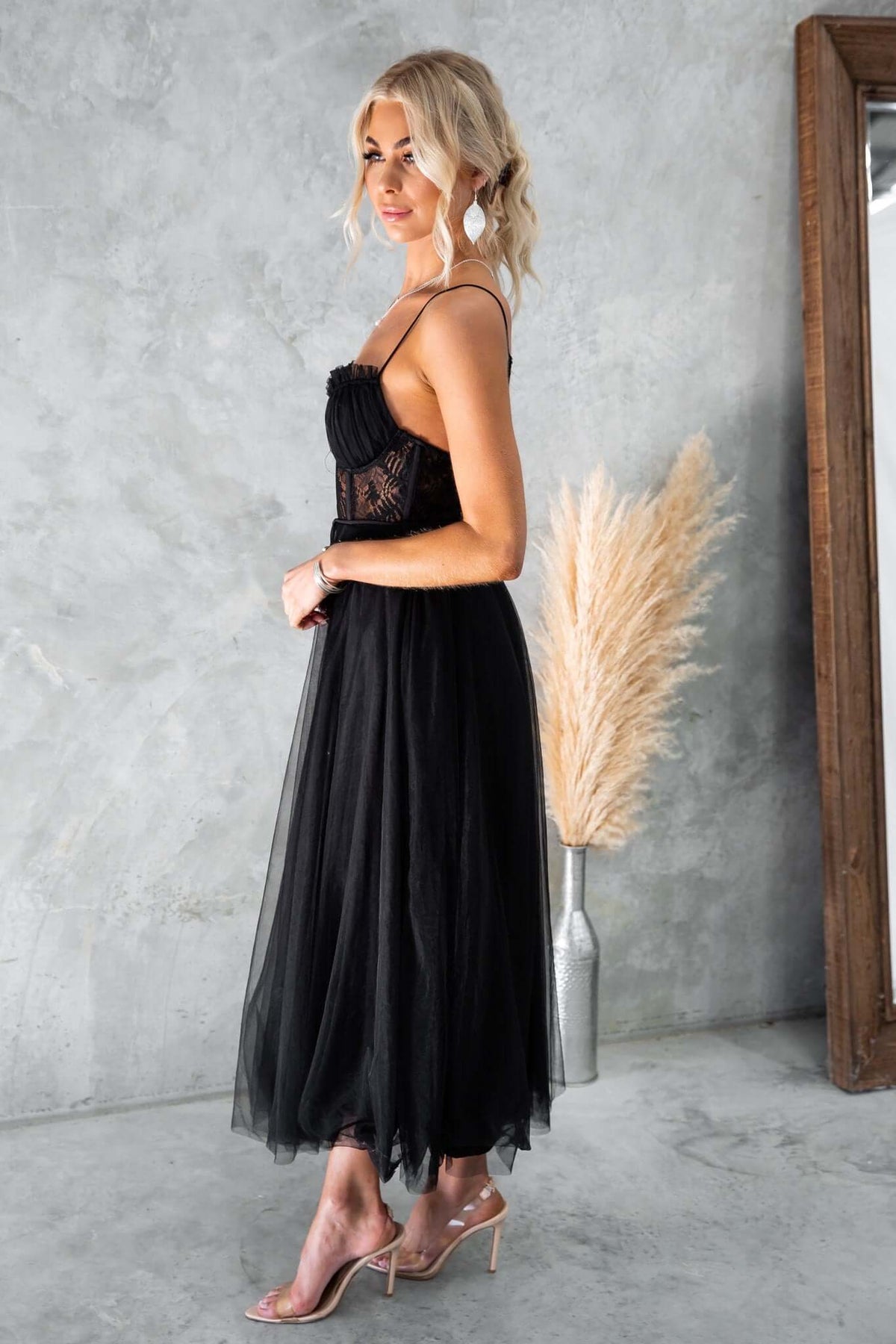 Model in a black tulle maxi dress with lace detail and adjustable straps, styled in a modern room.
