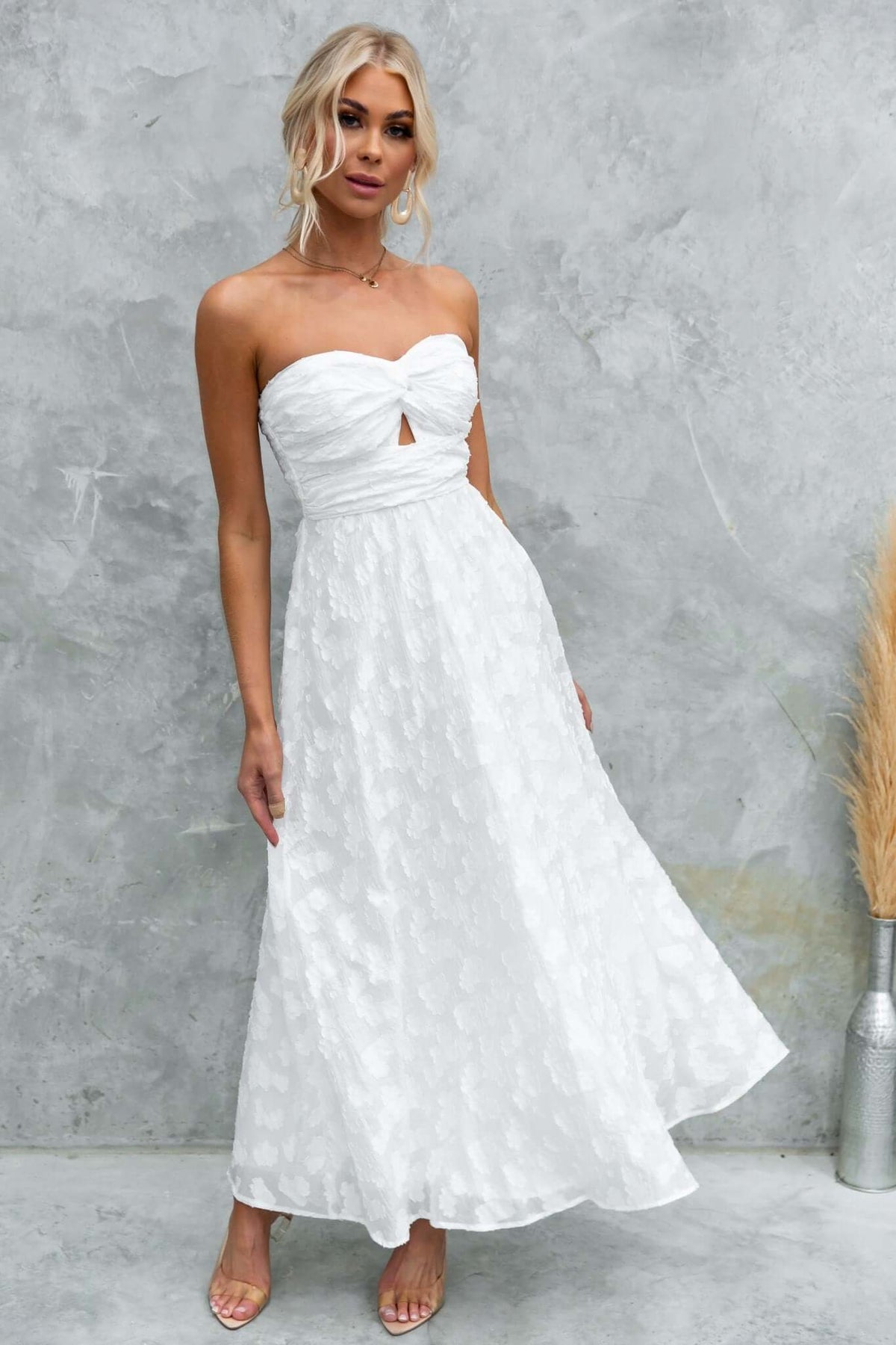 Model wearing the Ivrea Maxi Dress, a white strapless dress with a keyhole cutout and flowy skirt, set against a textured wall.