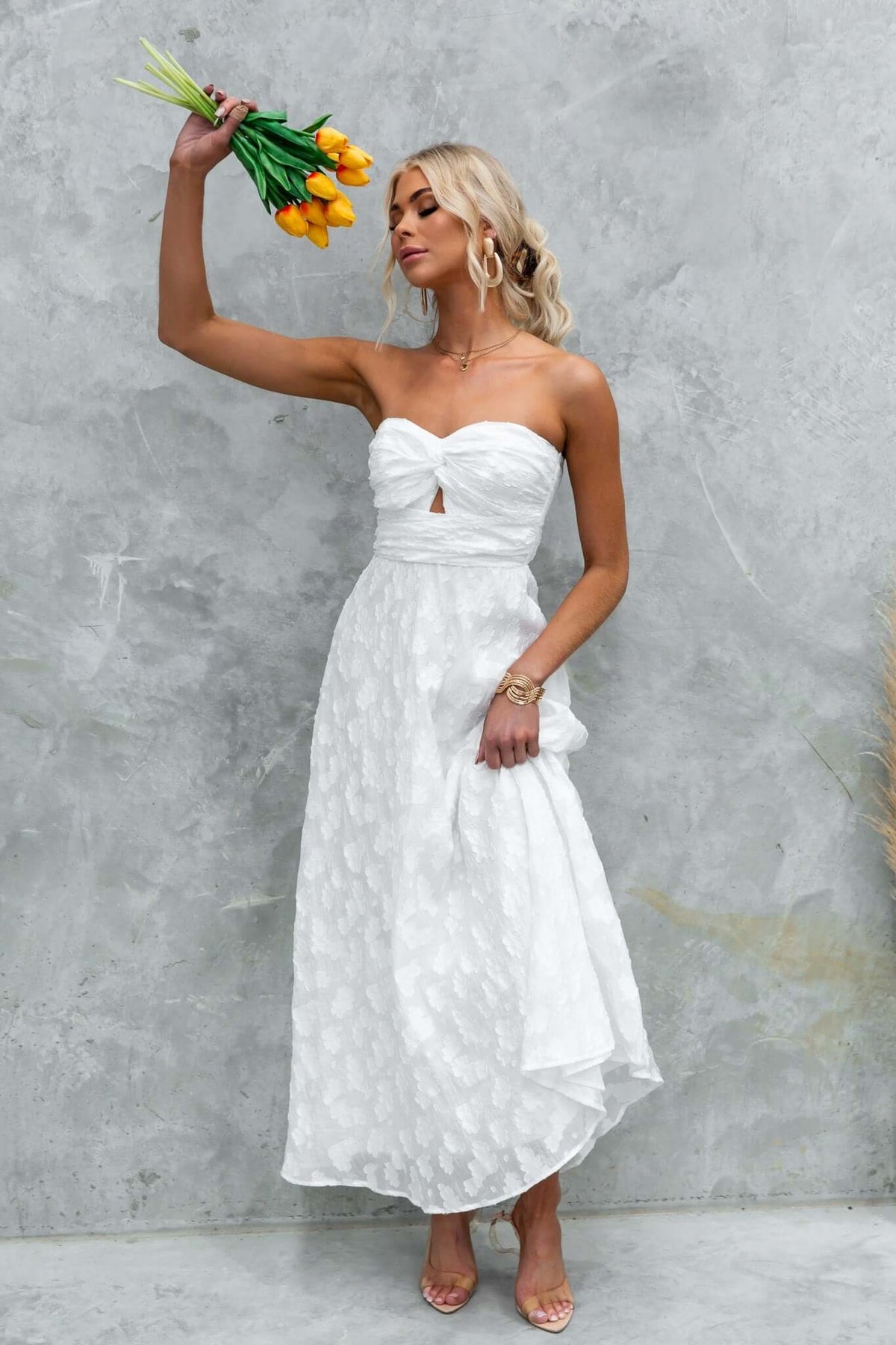 Model wearing Ivrea Maxi Dress, a strapless white dress with a knot detail, holding flowers against a textured background.