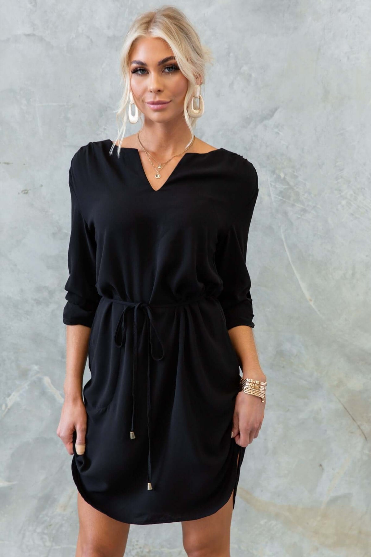 Model wearing the Deklyn Dress, a black V-neckline dress with a removable tie and pockets.