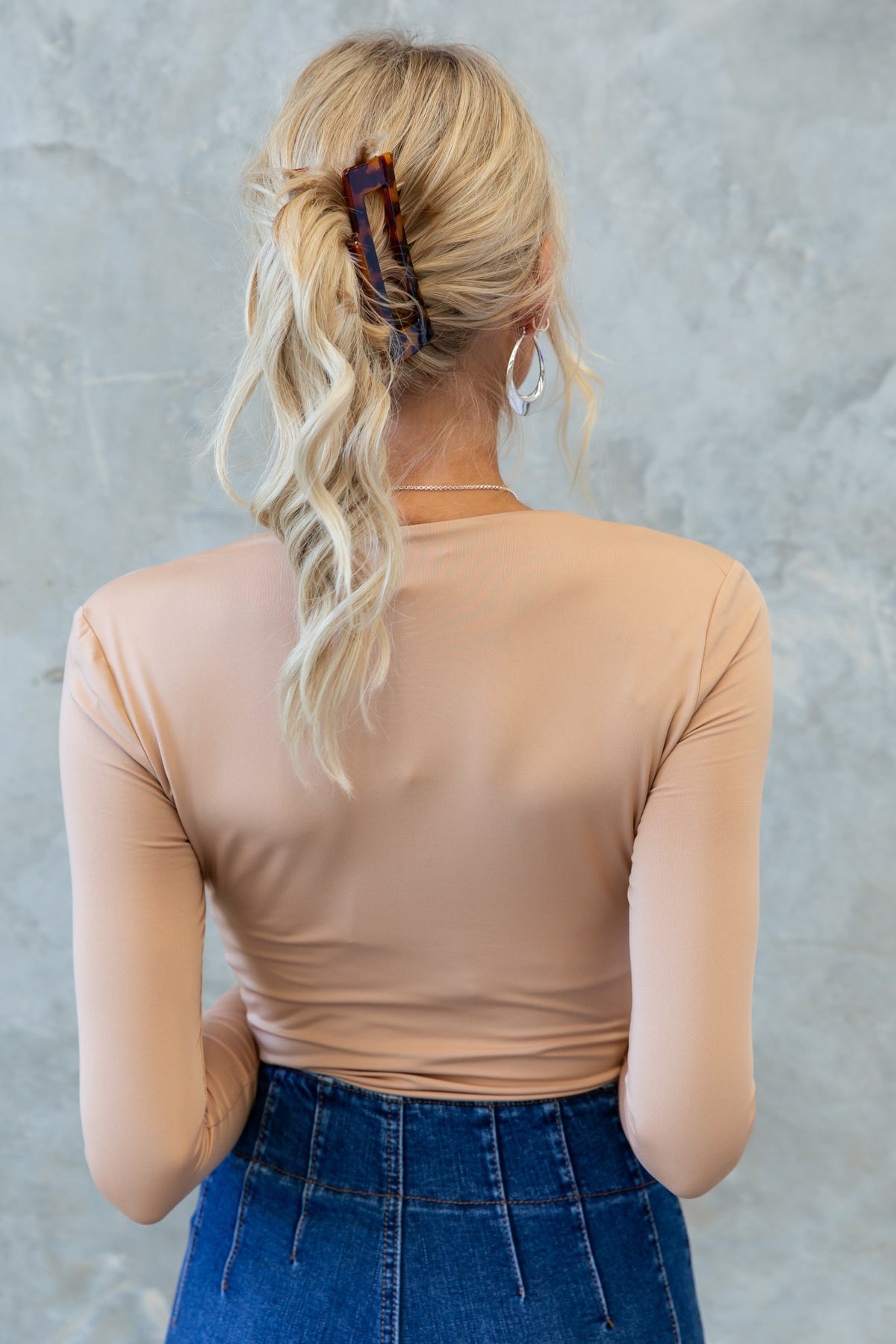 Back view of a model in a nude Unfair Bodysuit, featuring long sleeves and styled hair with an accessory.