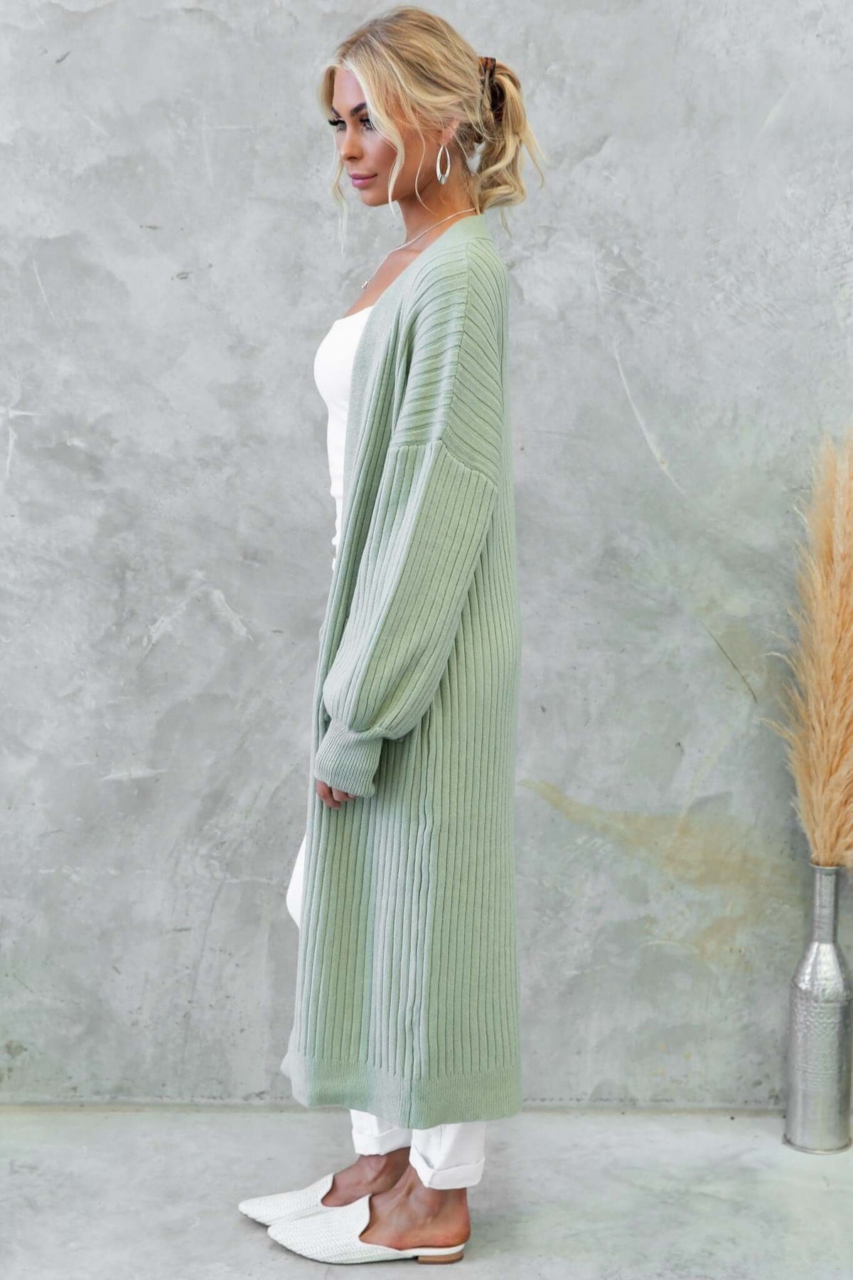 Side view of a woman wearing a longline, ribbed green Fano cardigan made from stretchy acrylic material.